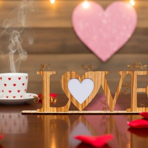 May include: A wooden "LOVE" sign with a heart-shaped cutout, a cup of coffee with heart designs, and scattered rose petals. A pink heart is in the background, with soft lighting.