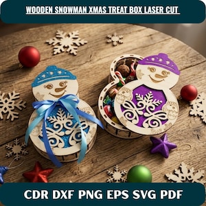 Puede incluir: Dos cajas de madera cortadas con láser en forma de muñeco de nieve. Una caja está cerrada con un inserto de purpurina azul y un lazo azul. La otra caja está abierta, revelando un inserto de purpurina morada y llena de pequeños dulces.