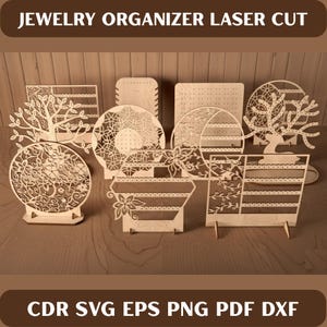 Peut inclure: Plusieurs présentoirs à bijoux en bois découpés au laser, de formes variées, dont des arbres, des cercles et des motifs géométriques. Les présentoirs sont exposés sur une surface en bois, avec le texte "JEWELRY ORGANIZER LASER CUT" en haut.