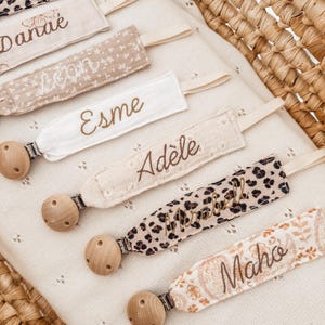 Peut inclure: Plusieurs attaches-tétines pour bébés avec des perles en bois et des lanières en tissu. Les lanières présentent divers motifs, dont des imprimés léopard et floraux, avec des noms brodés en fil doré. Les tétines reposent sur un tissu crème à pois.