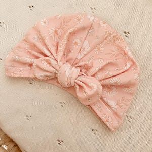 Puede incluir: Un turbante para bebé rosa claro con un estampado floral de flores y hojas blancas y rosa claro. El turbante está anudado en la parte delantera. El turbante está hecho de tela suave.