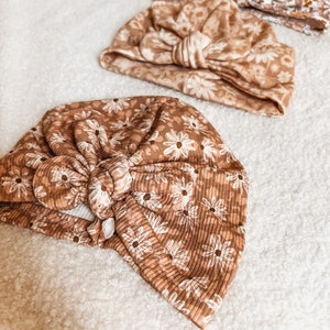 Puede incluir: Tres turbantes para bebés con estampados florales en tonos marrón y blanco. Los turbantes presentan un diseño anudado y están confeccionados en un tejido suave y acanalado. El estampado floral incluye pequeñas flores blancas en forma de margarita.