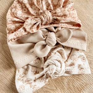 Może przedstawiać: Trzy opaski do włosów z tkaniny z kwiatowymi wzorami w odcieniach brązu i bieli. Opaski są zawiązane na górze w supeł i mają kokardkę z boku.