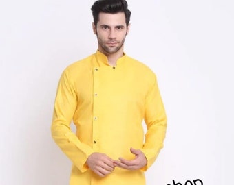 Puede incluir: Una camisa amarilla de manga larga con cuello mandarín y un solo botón de cierre en el lado izquierdo. La camisa es asimétrica, con el dobladillo más largo en el lado derecho. El modelo lleva pantalones blancos y sandalias marrones. El texto "Handmadekurtashop" es visible en la camisa.