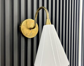 May include: A brass wall sconce with a white cone-shaped lampshade. The sconce is mounted on a black wall with vertical grooves.