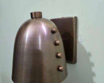 May include: A bronze-colored wall sconce with a bell-shaped shade and a square mounting plate. The shade has four decorative rivets. The fixture is mounted on a light green wall.
