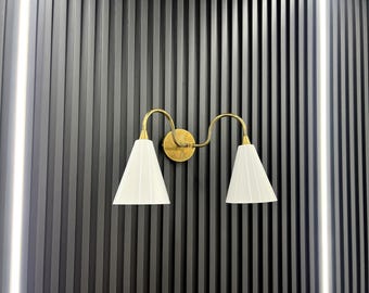 May include: A gold wall sconce with two white lampshades. The sconce is mounted on a black wall with vertical grooves.