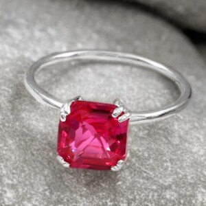 May include: A silver ring featuring a vibrant, square-cut, pink gemstone. The gemstone is held in place by four prongs, with a slender, polished band. The ring is presented on a textured grey surface.