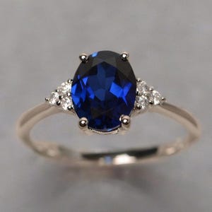 May include: A rose gold ring with a large, oval-cut, deep blue gemstone. Small, clear accent stones are set on either side of the main stone. The ring's band is a delicate, polished rose gold.