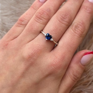 May include: A silver-toned ring featuring a central heart-shaped blue gemstone flanked by two small, clear round stones on a thin, polished metal band. The design is symmetrical, with the blue heart stone positioned as the primary focal point.