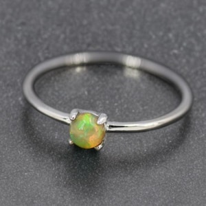 May include: A silver ring featuring a small, round opal gemstone. The opal has green, yellow, and orange tones. The ring's band is slender and polished, and the gemstone is held by four prongs.