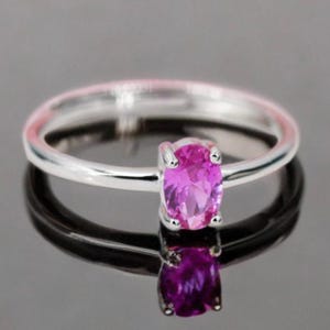 May include: A silver ring featuring a vivid pink oval gemstone. The gemstone is held in place by four prongs. The ring's band is slender and polished, reflecting light. A classic design.