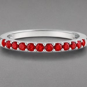 Op de afbeelding: Een zilverkleurige ring met een rij van twaalf kleine, ronde, felrode stenen die langs de voorste helft van de band zijn gezet. Het metaal heeft een gepolijste afwerking en de stenen worden op hun plaats gehouden door kleine, zilverkleurige pootjes.