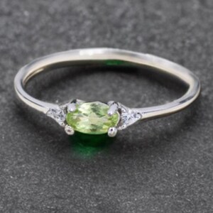 May include: A silver ring with a light green oval gemstone at its centre. The ring has small, clear accent stones either side of the main stone. The band is thin and polished, creating a delicate and elegant appearance.