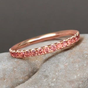 Può includere: Un sottile anello in oro rosa con una fila di piccole pietre preziose rotonde color arancio rosato incastonate lungo la metà superiore. Il metallo ha una finitura lucida e riflettente e le pietre sono di dimensioni uniformi.