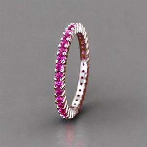 May include: A thin eternity band ring featuring a continuous row of small, round, vibrant pink gemstones set in a polished silver-toned metal setting. The ring is positioned vertically, showcasing the uniform circular arrangement of the stones.