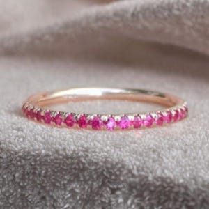 Può includere: Un sottile anello in oro rosa caratterizzato da una fila continua di piccole pietre preziose rotonde di colore rosa vivace incastonate lungo la metà anteriore della circonferenza metallica. L'anello ha una finitura lucida e un profilo sottile.