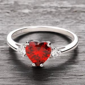 Puede incluir: Un anillo de plata con una piedra preciosa roja en forma de corazón sujeta por cuatro garras. La banda está adornada a cada lado con una piedra transparente más pequeña en forma de pera. El interior lleva grabado el texto S925 de plata.