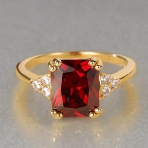 May include: A gold ring with a large, rectangular, deep red gemstone. Small, clear accent stones are set on either side of the main stone. The ring has a classic design, suitable for various occasions.