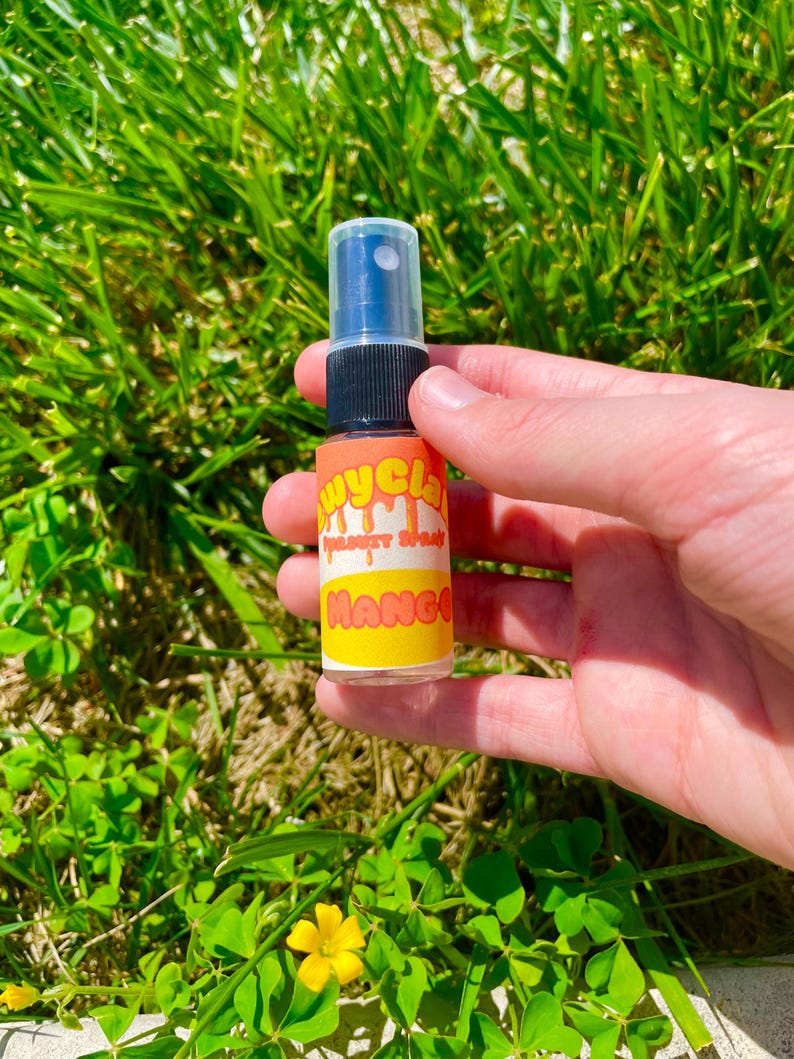 May include: A small clear plastic bottle with a black spray nozzle. The bottle has a yellow and orange label with the text "Juicy Glam" and "Mango" on it.