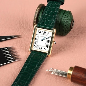 May include: A gold Cartier watch with a white face and Roman numeral hour markers. The watch has a green alligator leather strap.