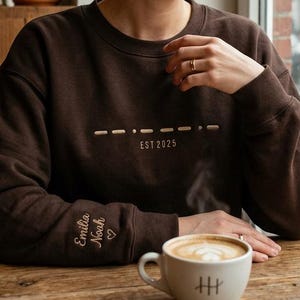 Könnte beinhalten: Ein braunes Sweatshirt mit dem Text "---- ---- EST 2025" auf der Brust. Der Ärmel hat die gestickten Namen "Emilia Noah" und ein Herz. Eine weiße Kaffeetasse mit einem Design steht auf einem Holztisch.