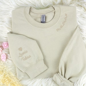 Könnte beinhalten: Ein hellbeiger Pullover mit den Worten "maumau" auf dem Kragen und "Leonie Fabius" mit einem Herz auf dem linken Ärmel gestickt.
