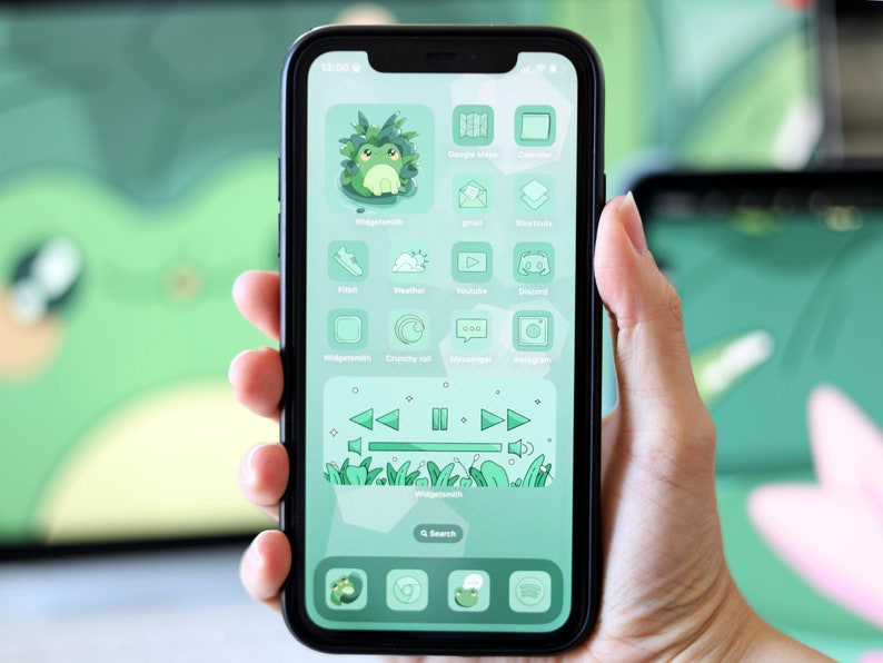 May include: A green and white iPhone with a custom home screen featuring a frog icon, a music player widget, and various app icons. The phone is being held in a hand.