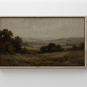 May include: A framed landscape painting in muted tones of brown, green, and gray. The artwork depicts a rural scene with rolling hills, trees, and a cloudy sky. The frame is a light wood color.
