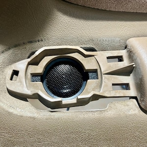 May include: A tan car door speaker with a black mesh cover. The speaker is set in a tan plastic housing with a rectangular shape and several openings. The speaker is part of the interior of a vehicle.