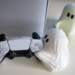 Pode incluir: Um controlador PlayStation 5 branco com botões pretos e um suporte em forma de fantasma branco. O suporte tem dois olhos pretos.