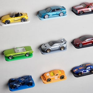 Op de afbeelding: Een verzameling van acht speelgoedauto's die op kleine planken worden getoond. De auto's zijn in verschillende kleuren, waaronder geel, groen, blauw, rood, zilver en zwart. De planken zijn bevestigd aan een witte muur.