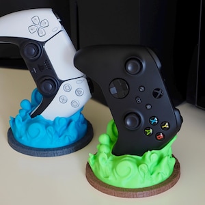 Könnte beinhalten: Zwei Videospielcontroller, einer weiß und einer schwarz, werden auf 3D-gedruckten Ständern präsentiert. Der weiße Controller befindet sich auf einem blauen Ständer mit Wellenmuster. Der schwarze Controller befindet sich auf einem grünen Ständer mit Wellenmuster.