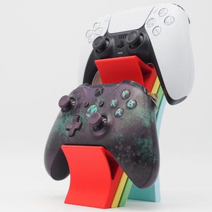 Op de afbeelding: Een veelkleurige gamecontrollerstandaard met een zwarte en blauwgroene Xbox-controller en een witte PlayStation 5-controller. De standaard heeft een rood, geel, groen en blauw ontwerp.