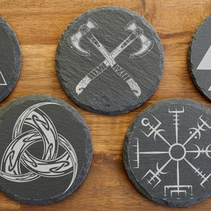Peut inclure: Ensemble de six sous-verres en ardoise noire avec différents motifs gravés, notamment un nœud celtique, une boussole, des haches croisées et un symbole Valknut.
