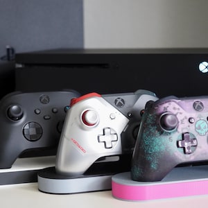Può includere: Quattro controller Xbox su supporti di ricarica. I controller sono neri, argentati, grigi e di un viola scuro con un design a forma di teschio. I supporti sono neri, grigi e rosa.