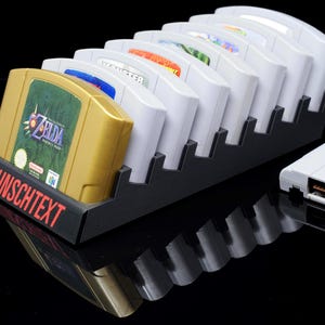 Op de afbeelding: Een zwarte, trapsgewijze displaystandaard met verschillende videospelcartridges. De voorste cartridge is goudkleurig met een groen label met de tekst "ZELDA". De basis van de standaard heeft het woord "WUNSCHTEXT" in het rood. Er staan ook verschillende witte cartridges in de standaard.