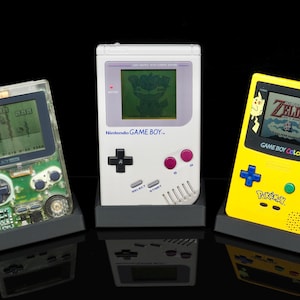 Op de afbeelding: Drie vintage Nintendo Game Boy-consoles. Een transparante, een witte en een gele Game Boy Color zijn te zien, elk met een spel op het scherm. De gele console heeft een Pokémon-ontwerp.