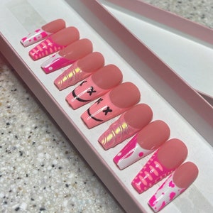 May include: A set of pink press-on nails with different designs. Some nails have a white base with pink and white swirls, others have a pink base with a black smiley face with an X over the eyes, and others have a pink base with a white and pink swirl design.