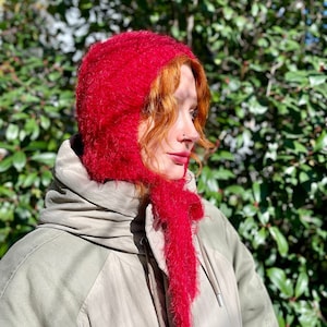 Op de afbeelding: Een vrouw draagt een felrode, pluche gebreide muts en een bijpassende sjaal. De muts is over haar oren getrokken en de sjaal is losjes om haar nek geknoopt.