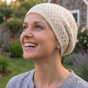 May include: A cream-colored crochet beanie featuring a textured, repeating shell stitch pattern. The hat is constructed from soft yarn and fits snugly around the head, providing full coverage with a slightly slouchy silhouette at the back.