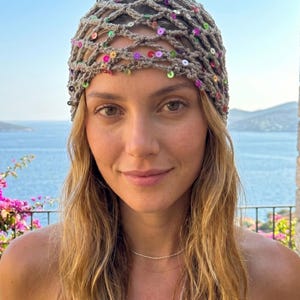 Peut inclure: Un couvre-chef en crochet avec un motif en filet, orné de sequins colorés. Le couvre-chef est de couleur beige neutre, avec des sequins dans les tons de rose, vert et or. L'accessoire est porté à l'extérieur.