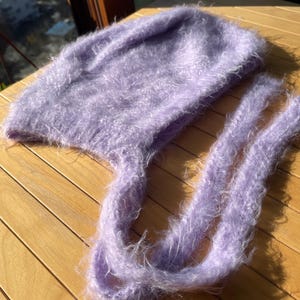 A soft, lavender fuzzy knitted bonnet lying on a light-colored wooden table. The bonnet features long matching ties and a fluffy, mohair-style texture. Bright, natural sunlight streams across the scene, highlighting the delicate fibers of the knitwear.