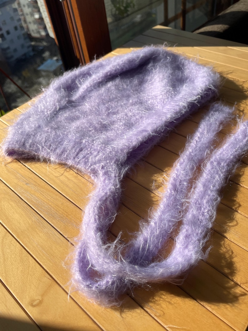 A soft, lavender fuzzy knitted bonnet lying on a light-colored wooden table. The bonnet features long matching ties and a fluffy, mohair-style texture. Bright, natural sunlight streams across the scene, highlighting the delicate fibers of the knitwear.