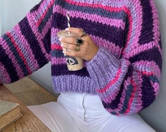 Puede incluir: Un suéter de punto grueso a rayas en tonos morados, rosas y grises. El suéter tiene cuello redondo y corte cropped. Las mangas son largas. La persona lleva vaqueros blancos. El suéter está hecho de lana gruesa.