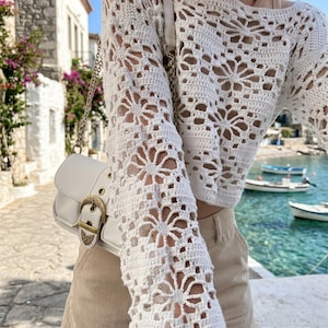 May include: A white crochet top with long sleeves featuring a floral pattern. The top is paired with beige shorts and a white purse with a gold buckle. The image is taken outdoors with a scenic background.