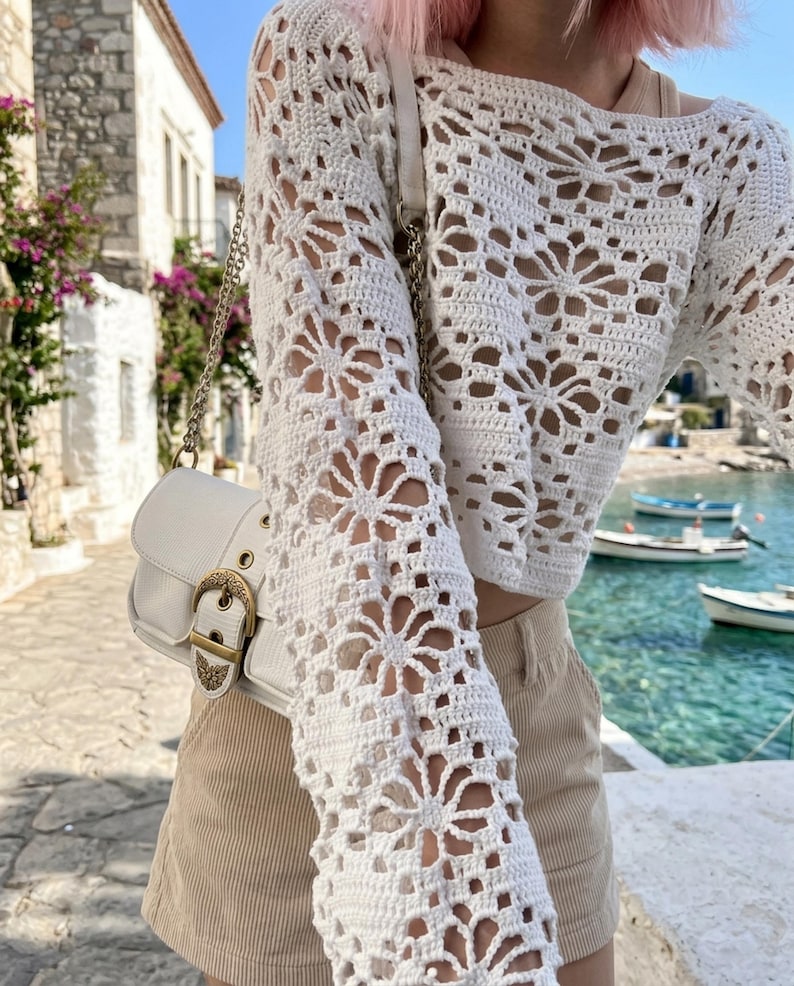 May include: A white crochet top with long sleeves featuring a floral pattern. The top is paired with beige shorts and a white purse with a gold buckle. The image is taken outdoors with a scenic background.