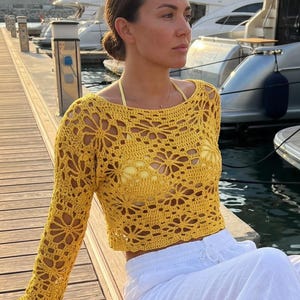 A woman sitting by a harbor wearing a bright yellow handmade crochet long-sleeve crop top with intricate lace patterns and white linen trousers.