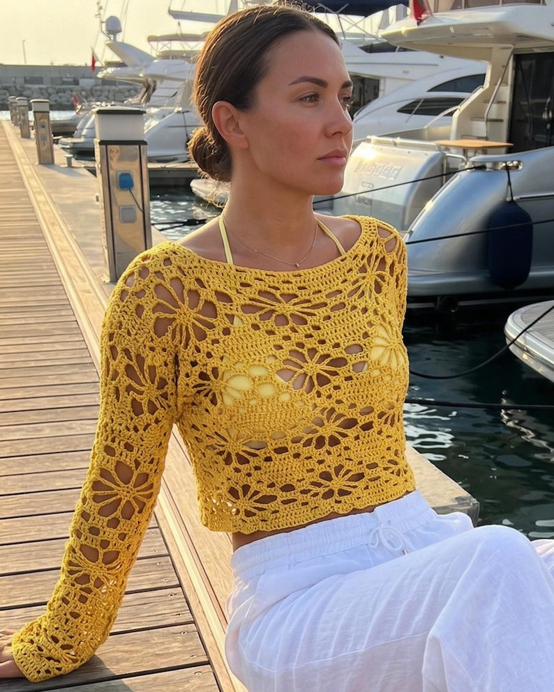 A woman sitting by a harbor wearing a bright yellow handmade crochet long-sleeve crop top with intricate lace patterns and white linen trousers.