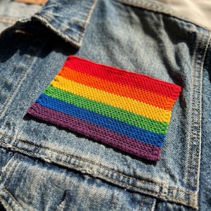May include: A rainbow-colored, rectangular patch with red, orange, yellow, green, blue, and purple stripes. The patch is made of a textured material and is placed on a denim jacket.